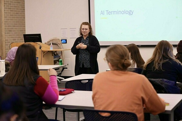 A woman with light brown curly hair and glasses, wearing a black cardigan and polka dot shirt, speaks to students in a classroom. "AI Terminology" is projected on the screen. Students in orange and pink tops listen.